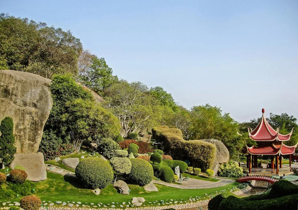 Landscaped garden with large boulders, clipped shrubs and a red pagoda beside a stone path under a clear sky