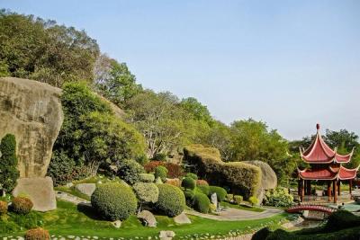 Landscaped garden with large boulders, clipped shrubs and a red pagoda beside a stone path under a clear sky