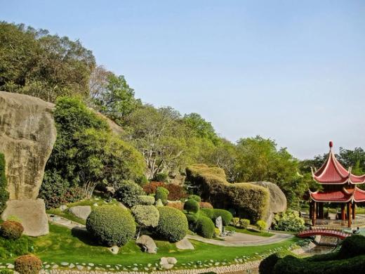Landscaped garden with rock formations, clipped shrubs, trees and a red pagoda beside a curving path under a clear sky.