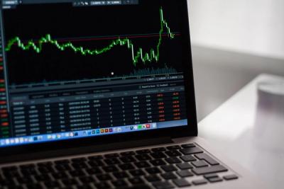 Laptop screen showing a green candlestick trading chart and a table of trade data on a desk.