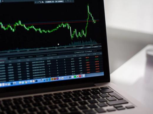 Laptop screen showing a green candlestick trading chart and a table of trade data on a desk.