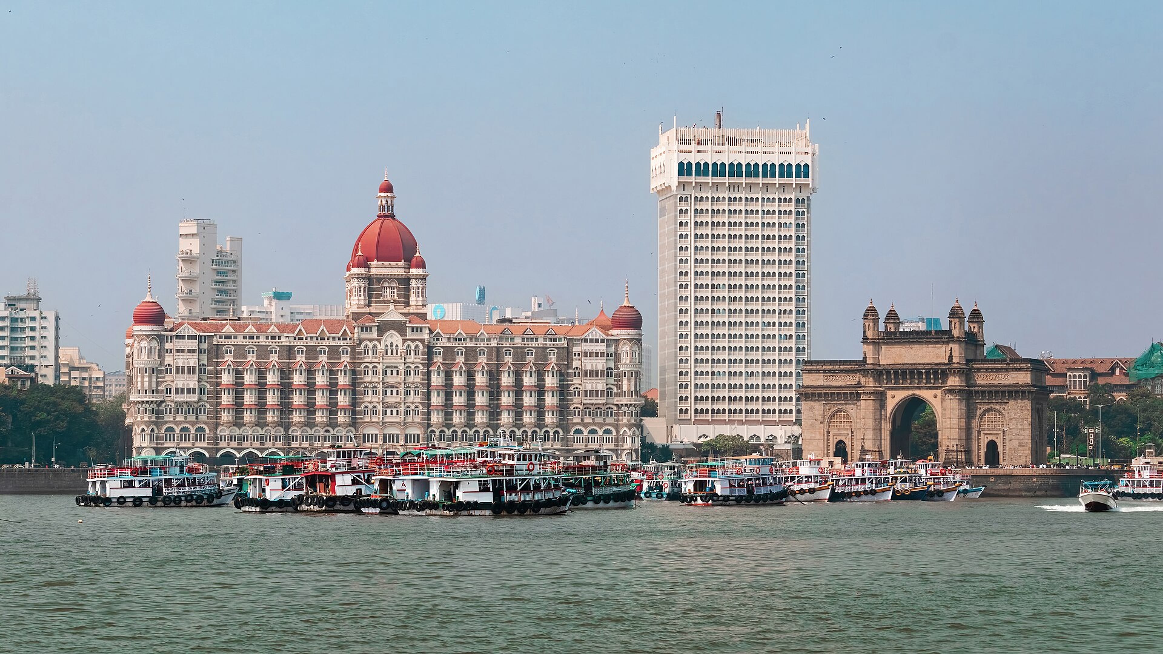 Waterfront view of a domed heritage hotel and a tall white tower beside an arched stone gateway, with boats moored in the foreground