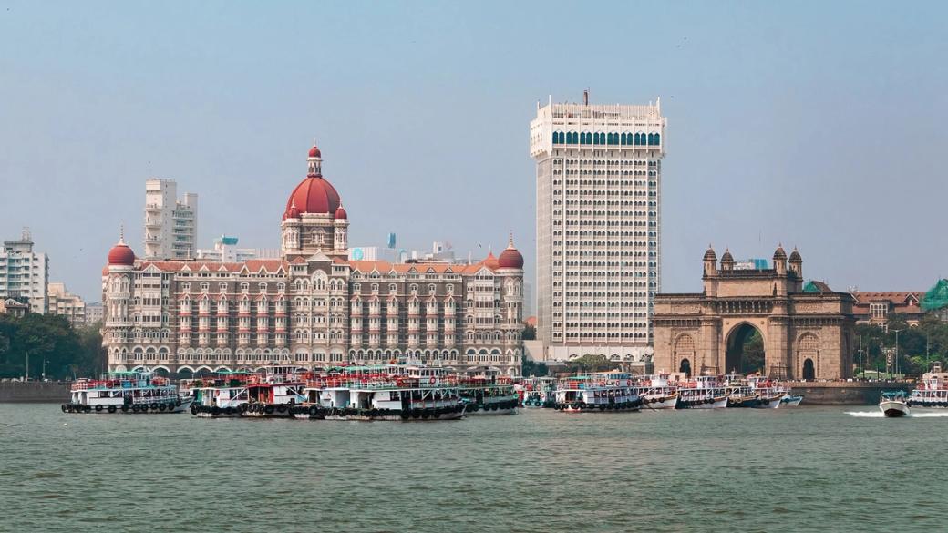 Waterfront view of a domed heritage hotel and a tall white tower beside an arched stone gateway, with boats moored in the foreground