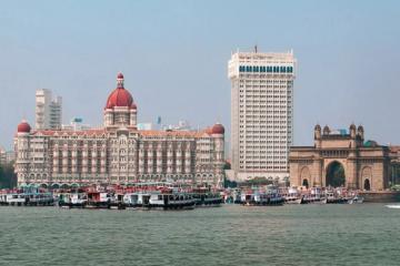 Waterfront view of a domed heritage hotel and a tall white tower beside an arched stone gateway, with boats moored in the foreground