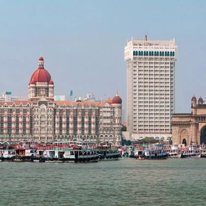Waterfront view of a domed heritage hotel and a tall white tower beside an arched stone gateway, with boats moored in the foreground