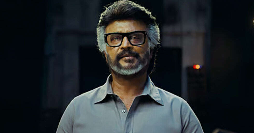 Man with grey hair and beard, wearing black-framed glasses and a grey shirt, looks ahead against a dark background