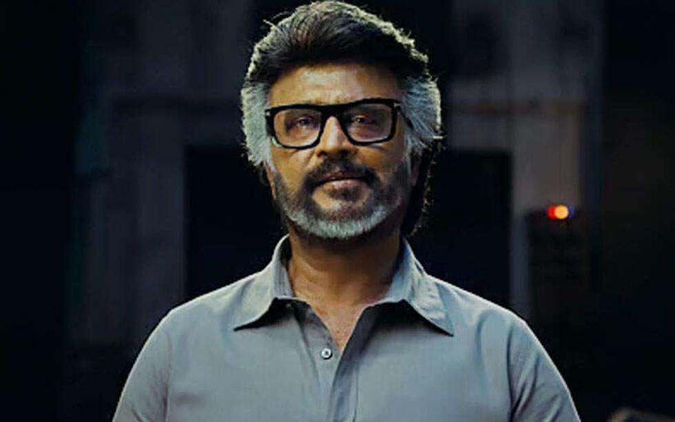 Man with grey hair and beard, wearing black-framed glasses and a grey shirt, looks ahead against a dark background