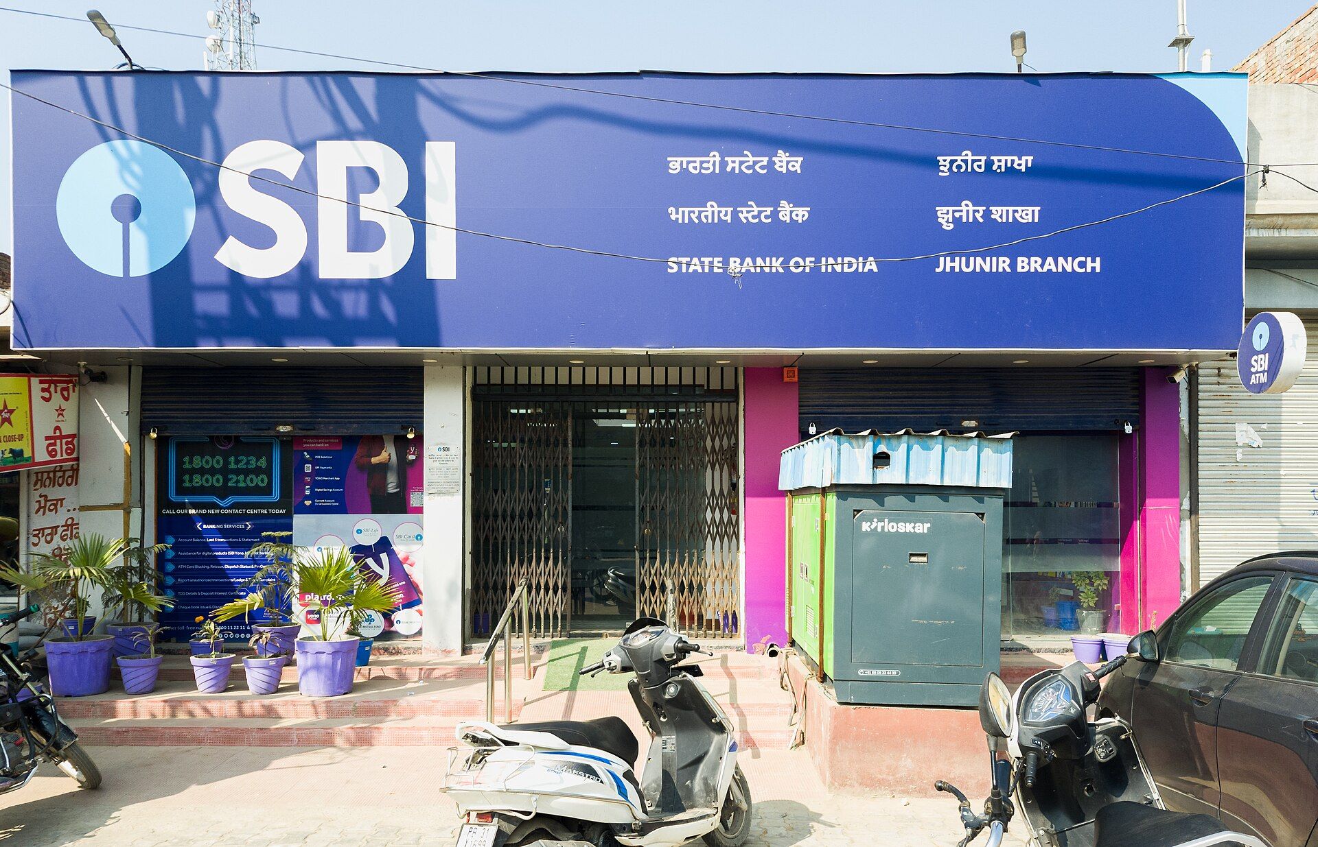 Front of a State Bank of India branch with a blue SBI signboard, a kiosk, parked scooters and potted plants