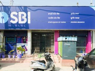 Front of a State Bank of India branch with a blue SBI signboard, a kiosk, parked scooters and potted plants
