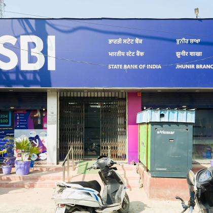 Front of a State Bank of India branch with a blue SBI signboard, a kiosk, parked scooters and potted plants
