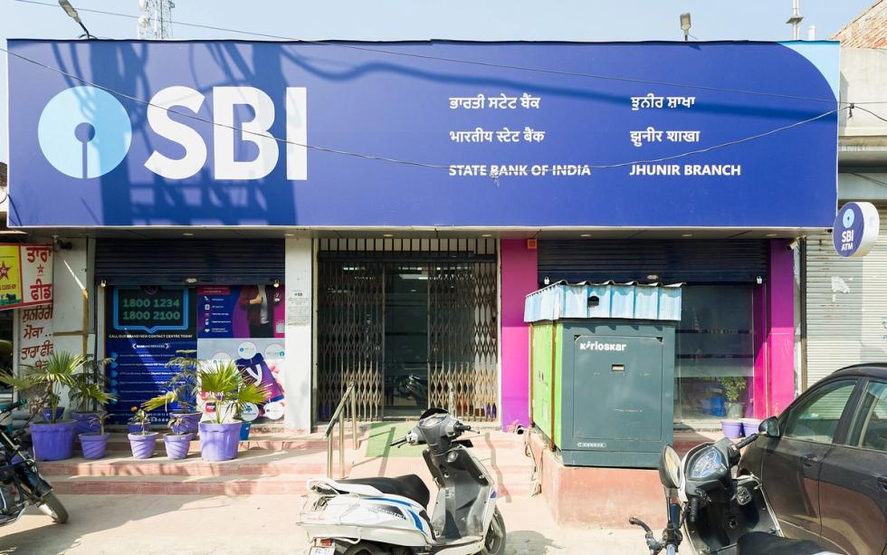 Front of a State Bank of India branch with a blue SBI signboard, a kiosk, parked scooters and potted plants