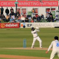 A batter in white kit and helmet plays a shot on a green field, a fielder in the foreground and spectators behind