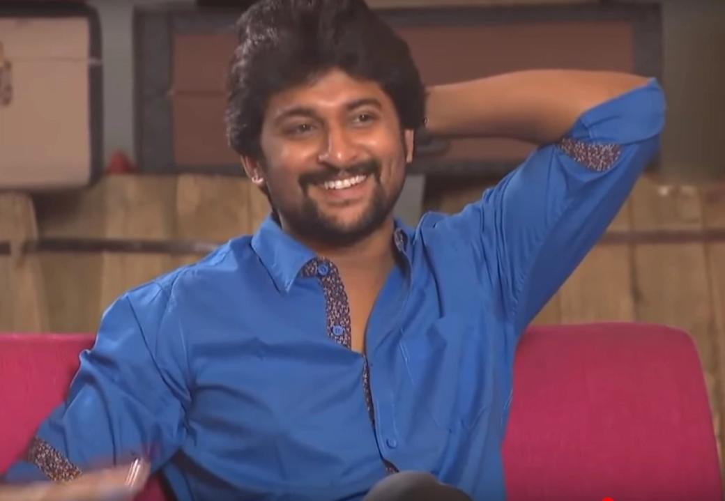 Smiling man in a blue shirt with one arm raised behind his head, seated on a pink sofa during a studio interview.