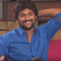 Smiling man in a blue shirt with one arm raised behind his head, seated on a pink sofa during a studio interview.