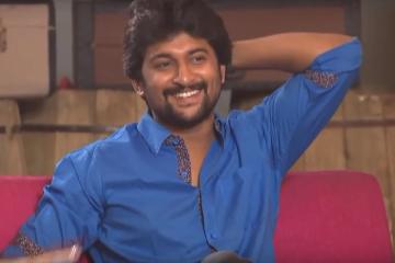 Smiling man in a blue shirt with one arm raised behind his head, seated on a pink sofa during a studio interview.