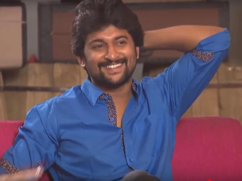 Smiling man in a blue shirt with one arm raised behind his head, seated on a pink sofa during a studio interview.