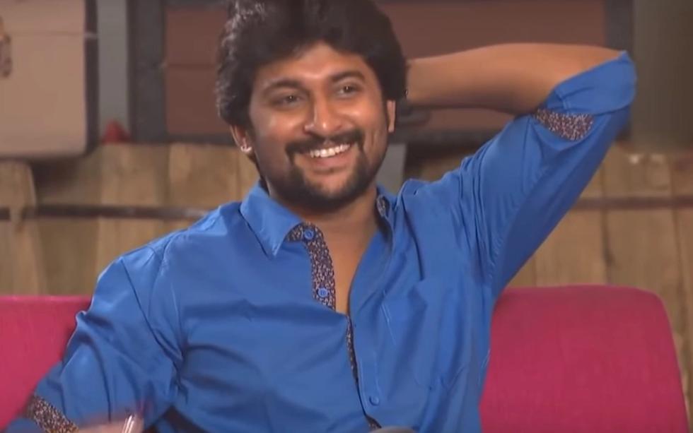 Smiling man in a blue shirt with one arm raised behind his head, seated on a pink sofa during a studio interview.
