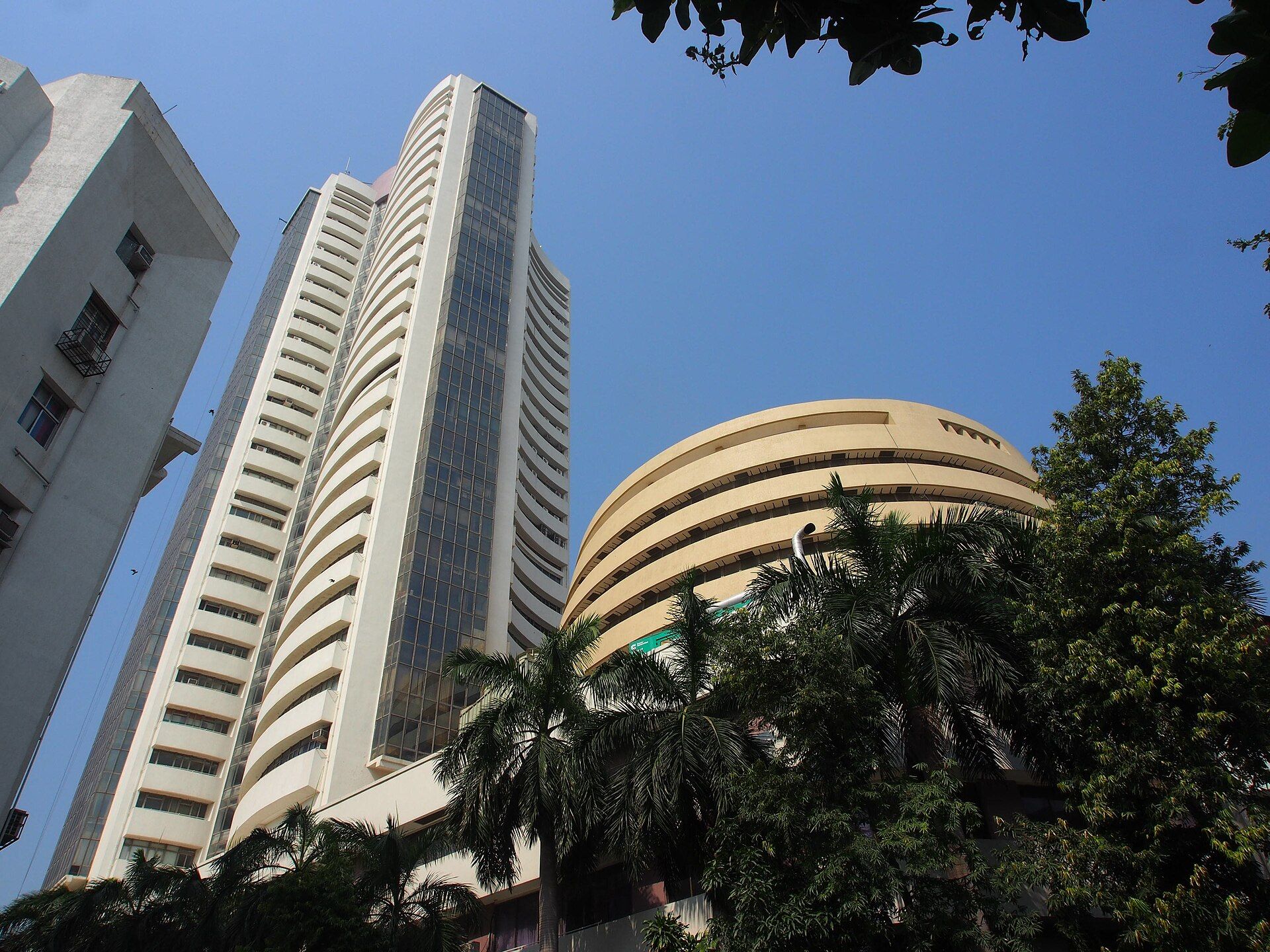 Curved cream-coloured high-rise and a round building seen from below, with palm trees and a blue sky