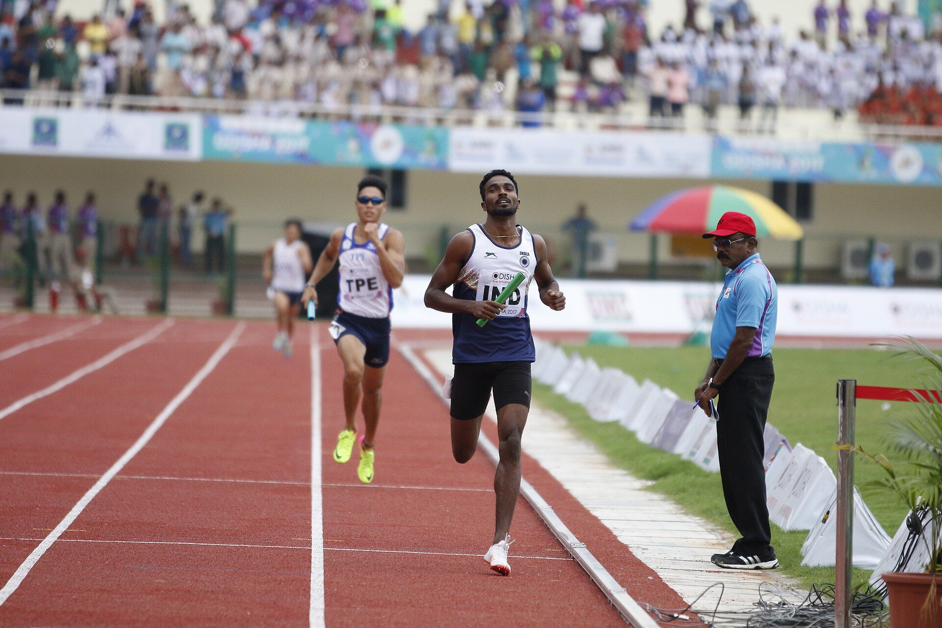 Two relay runners, one carrying a baton, race along a red track as an official in a blue shirt watches from the side
