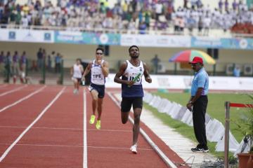 Two relay runners, one carrying a baton, race along a red track as an official in a blue shirt watches from the side