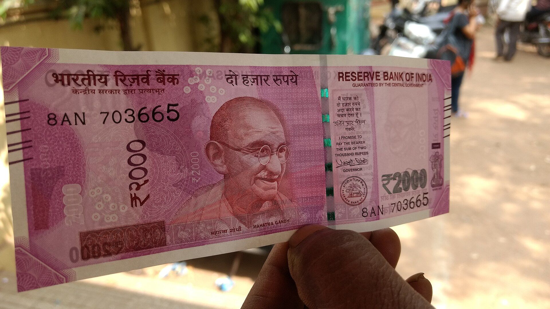 A hand holding a ₹2000 banknote up in front of a blurred street with people and motorbikes