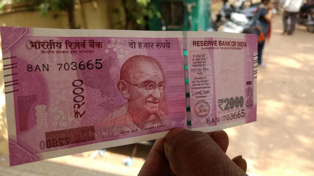 A hand holding a ₹2000 banknote up in front of a blurred street with people and motorbikes