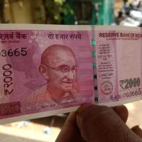 A hand holding a ₹2000 banknote up in front of a blurred street with people and motorbikes