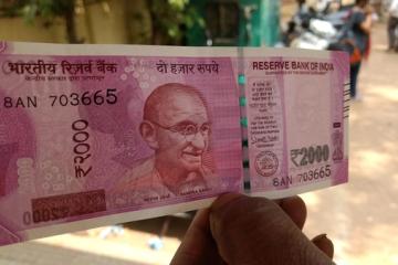 A hand holding a ₹2000 banknote up in front of a blurred street with people and motorbikes