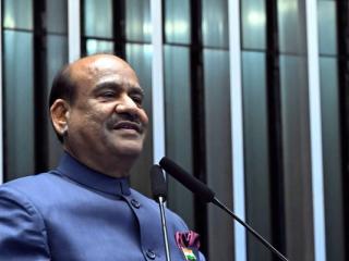 Man in a blue bandhgala jacket speaks at a wooden lectern with an India placard and microphones.