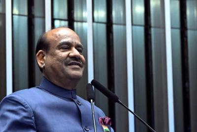 Man in a blue bandhgala jacket speaks at a wooden lectern with an India placard and microphones.