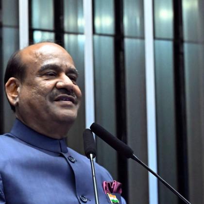 Man in a blue bandhgala jacket speaks at a wooden lectern with an India placard and microphones.