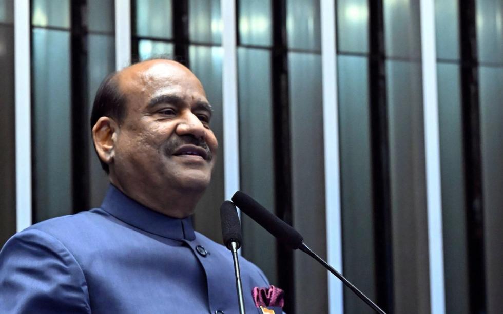 Man in a blue bandhgala jacket speaks at a wooden lectern with an India placard and microphones.