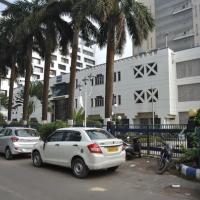 White multi-storey building with a red-tiled entrance canopy, palm trees, people at the gate and parked cars in front.