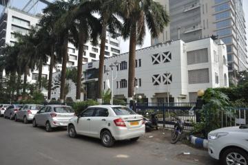 White multi-storey building with a red-tiled entrance canopy, palm trees, people at the gate and parked cars in front.