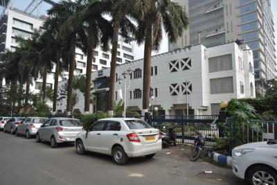 White multi-storey building with a red-tiled entrance canopy, palm trees, people at the gate and parked cars in front.
