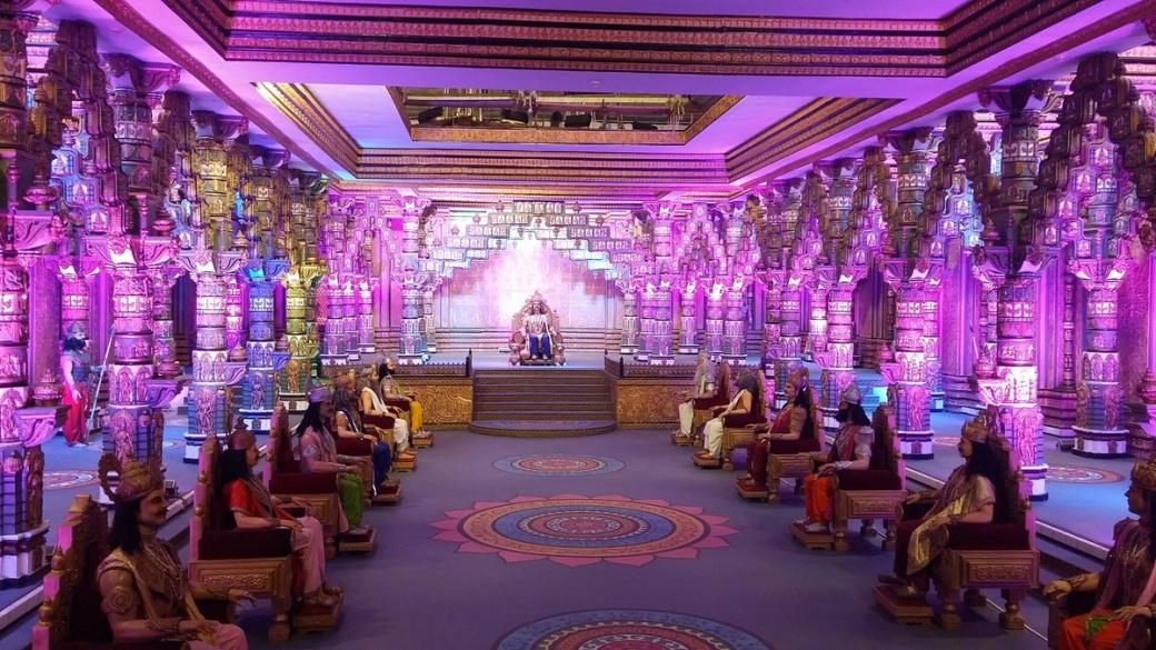 Ornate pillared hall lit in purple, with seated figures in traditional dress on a raised platform