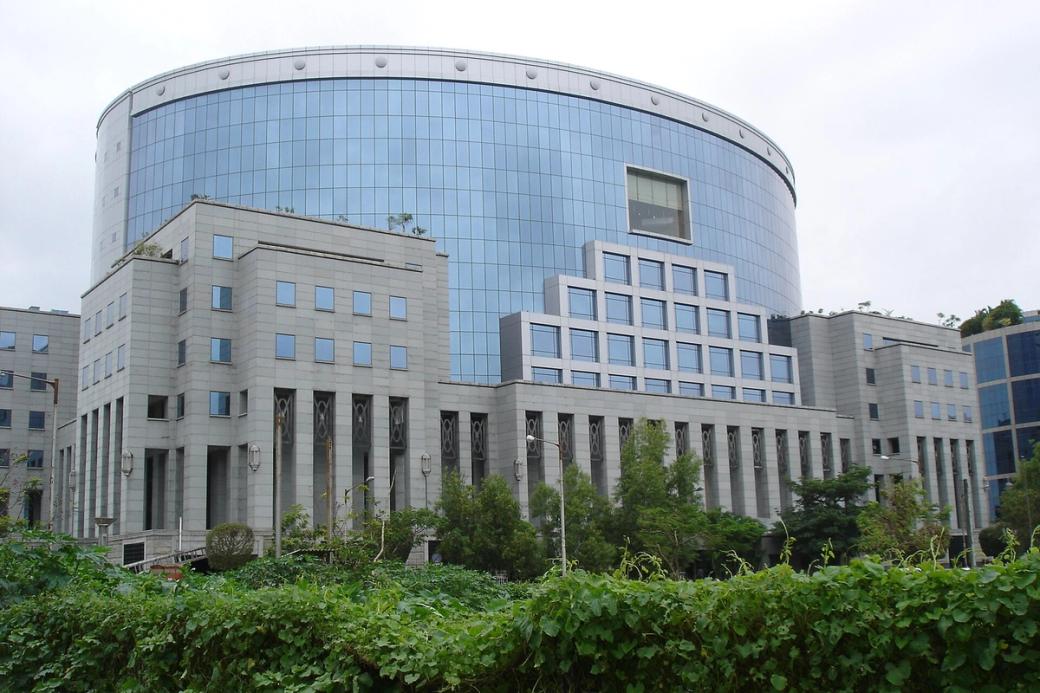 Curved glass office tower with a stone-clad base and green hedge under an overcast sky, Bandra-Kurla Complex.