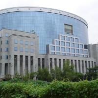 Curved glass office tower with a stone-clad base and green hedge under an overcast sky, Bandra-Kurla Complex.