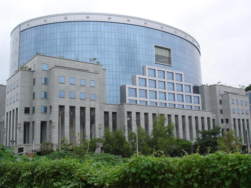 Curved glass office tower with a stone-clad base and green hedge under an overcast sky, Bandra-Kurla Complex.