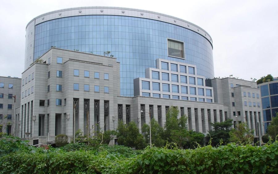 Curved glass office tower with a stone-clad base and green hedge under an overcast sky, Bandra-Kurla Complex.