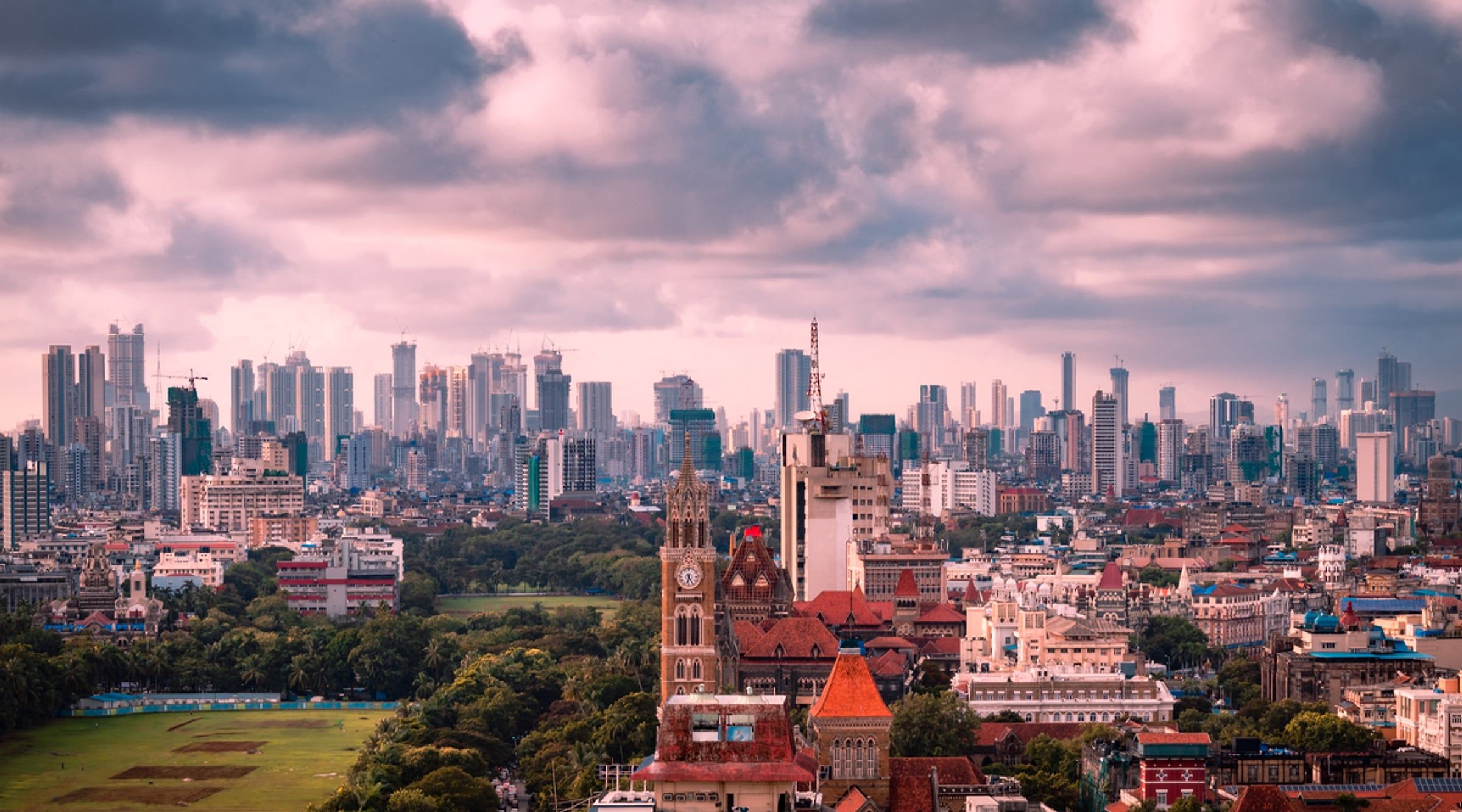Mumbai skyline of high-rise towers, a clock tower and green parkland under a cloudy sky
