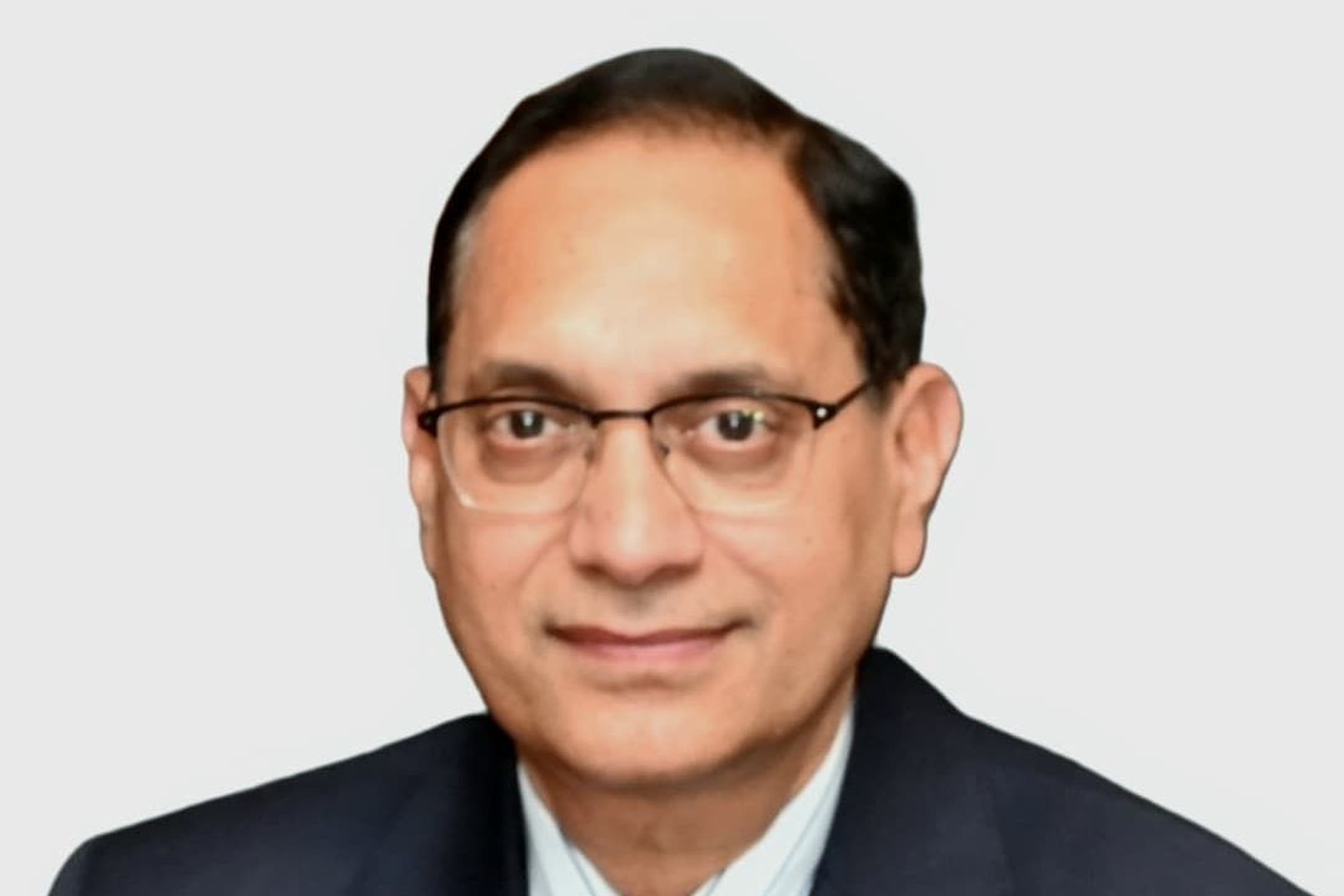 Man in dark suit, blue tie and glasses, smiling slightly against a plain light grey background