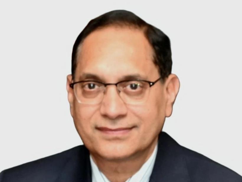 Man in dark suit, blue tie and glasses, smiling slightly against a plain light grey background