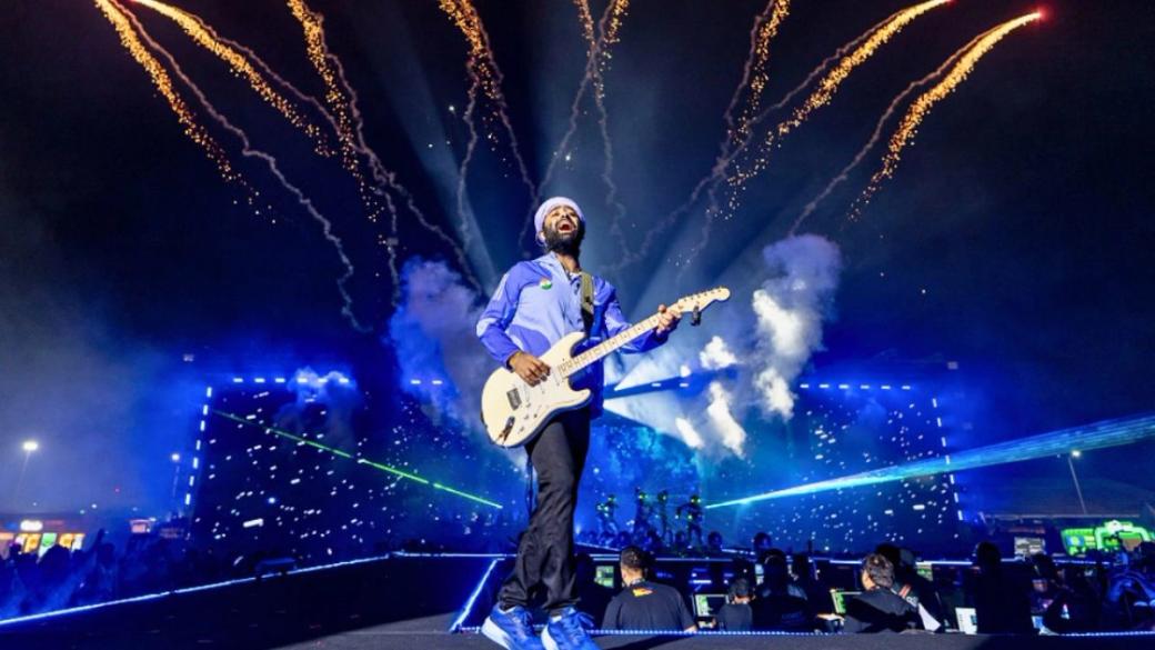 Arijit Singh playing an electric guitar and singing on a night stage, with fireworks and blue lights behind him.