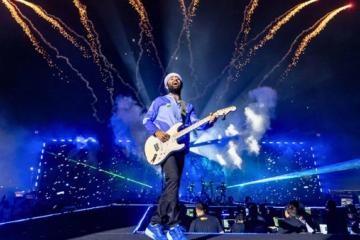 Arijit Singh playing an electric guitar and singing on a night stage, with fireworks and blue lights behind him.