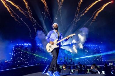 Arijit Singh playing an electric guitar and singing on a night stage, with fireworks and blue lights behind him.