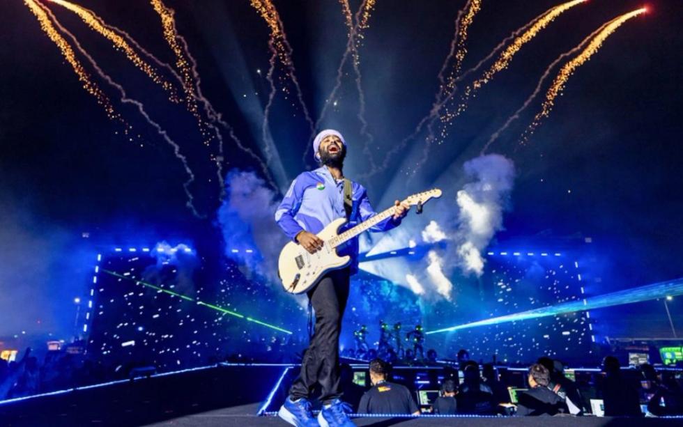 Arijit Singh playing an electric guitar and singing on a night stage, with fireworks and blue lights behind him.