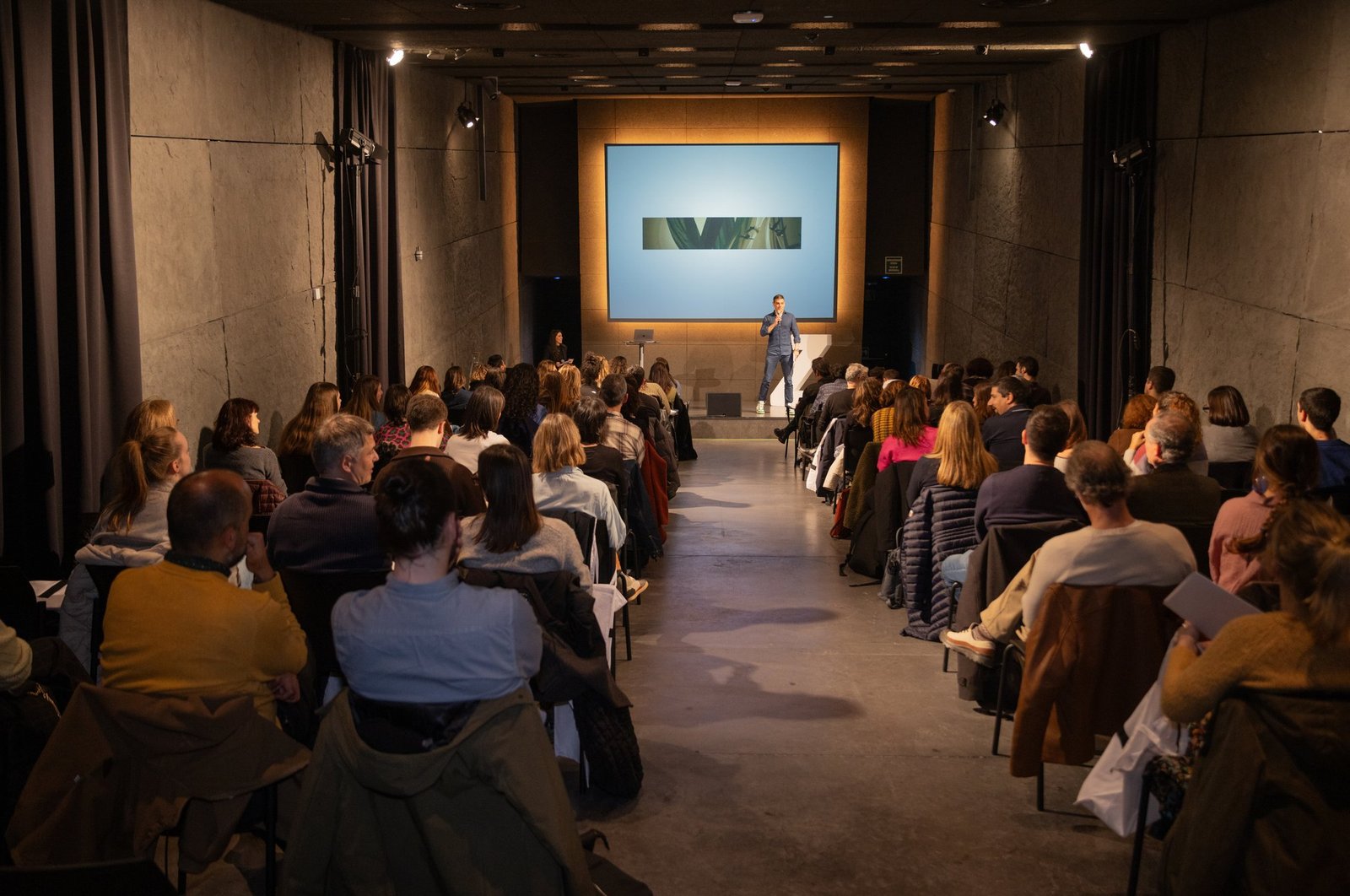 Packed room facing a speaker with a screen in the background