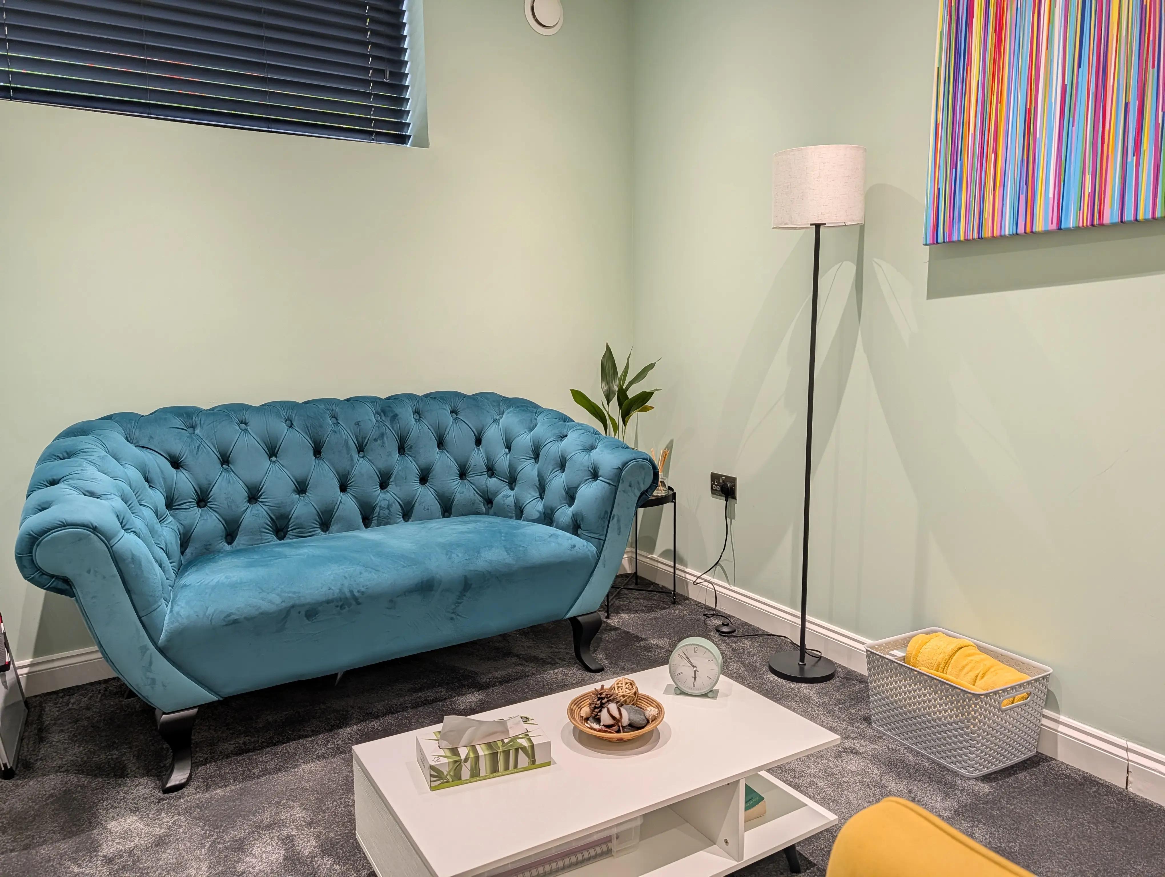 A therapy room used for in-person sessions in Chelmsford, with a teal velvet sofa, a low table with tissues, a floor lamp and a striped canvas on a pale green wall