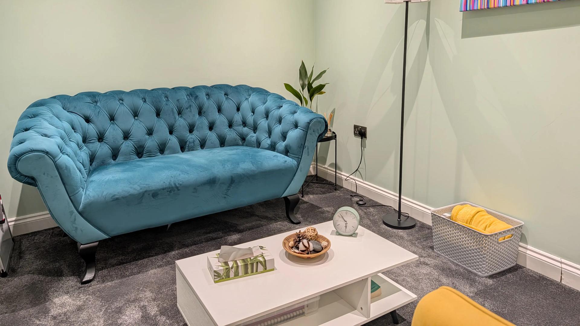 A therapy room used for in-person sessions in Chelmsford, with a teal velvet sofa, a low table with tissues, a floor lamp and a striped canvas on a pale green wall